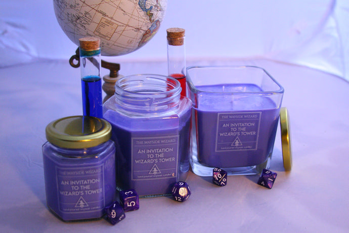All of the Wayside Wizard's sizes of the themed dnd candle - An invitation to the Wizard's Tower. Dice are scattered around, a globe is in the background and a few vials that resemble potions are behind the candles. 